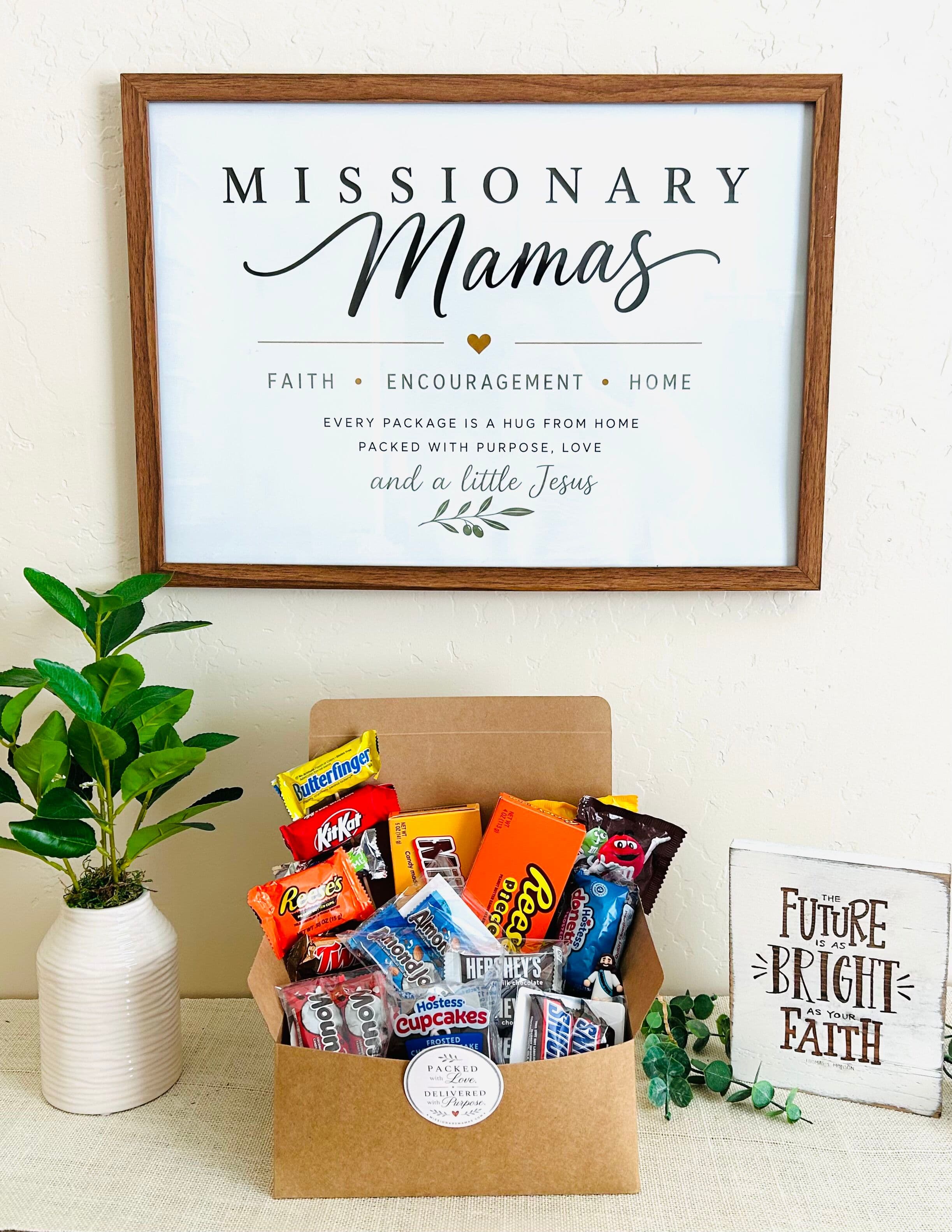 A Missionary Mamas care box filled with candy and treats beneath a framed 'Faith, Encouragement, Home' sign